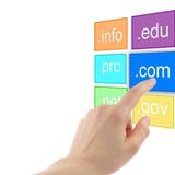 Alt text: Hand pointing at ".com" domain on colorful domain extension blocks.
