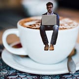 Miniature person with laptop sitting on coffee cup.