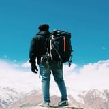 Backpacker standing on mountain peak with snowy slopes and blue sky, enjoying outdoor adventure.