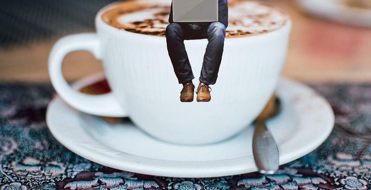 Miniature person with laptop sitting on coffee cup.