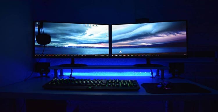 Computer desk with dual screens, gaming keyboard, and ambient blue LED backlit setup.