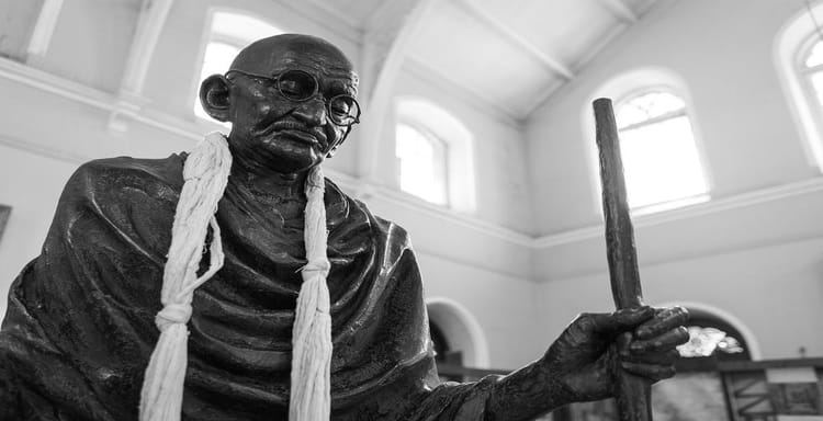 Mahatma Gandhi statue holding a walking stick, located inside a museum with large windows and high ceilings.