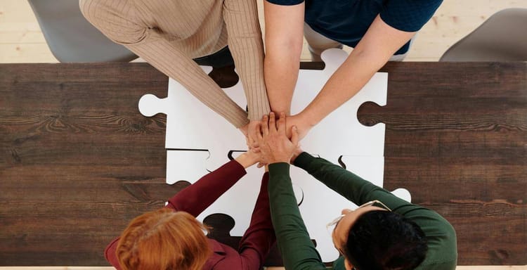 Alt text: Three people join hands over a puzzle piece on a wooden table, symbolizing teamwork and collaboration.
