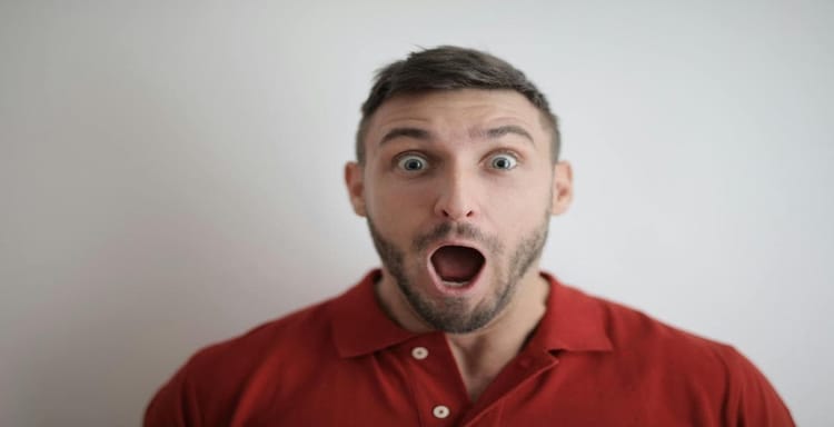Alt text: Young man with surprised expression facing camera, wearing red shirt, neutral background.