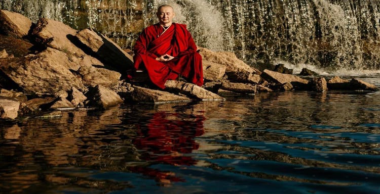 Meditating monk in red robes near waterfall with calm water reflection.