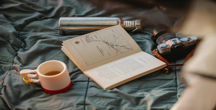 Portable camera, book, cup, and flashlight on bed for writing and reading.