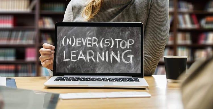 Alt text: Woman holding a laptop with a blackboard displaying (N)ever (S)top Learning in a library.