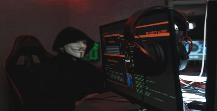 A hacker in a dark hoodie with Guy Fawkes mask working on cybersecurity, surrounded by monitors displaying security alerts and hacking data.
