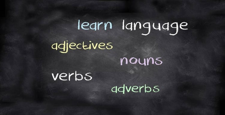 Alt text: Image showing a blackboard with colorful words related to language learning and writing skills like nouns, verbs, adjectives, and adverbs.