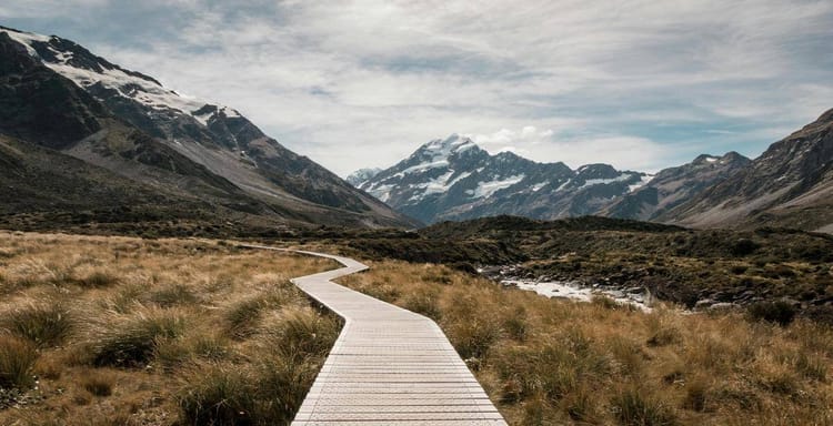 Scenic mountain landscape with wooden walkway and snow-capped peaks in the background.