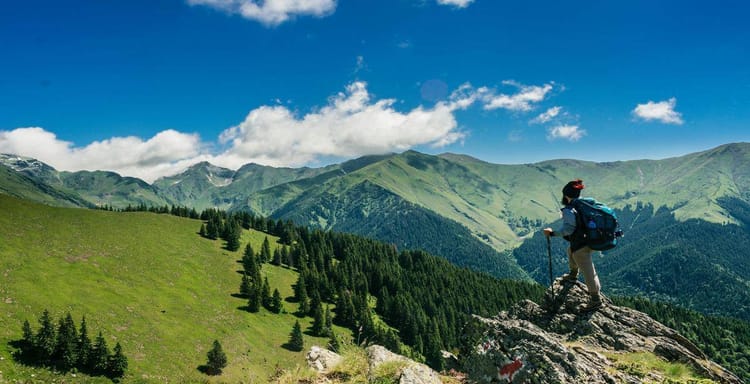 Hiker with backpack on rocky mountain ledge overlooking lush green hills and mountain range under blue sky.