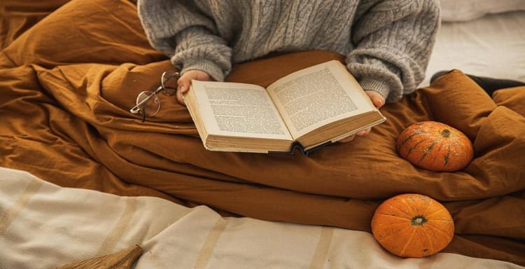 Close-up of person reading a book on bed with pumpkins for autumn vibe.