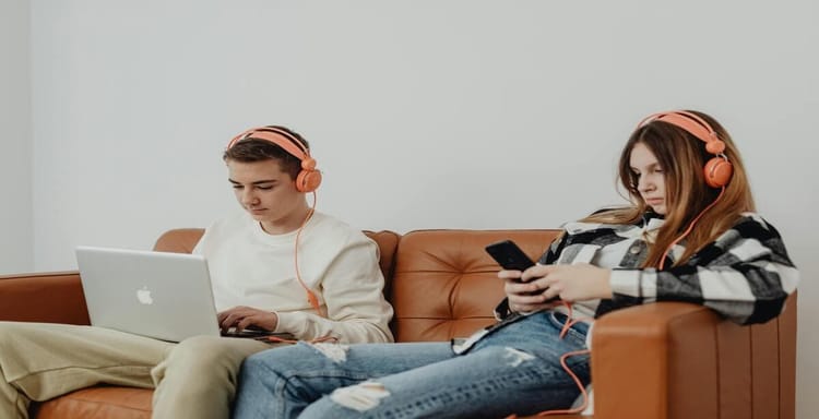 Alt text: Two teenagers with headphones using laptop and smartphone on brown couch, focusing on word and character counting.