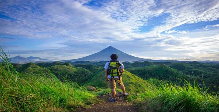 Hiker standing on trail overlooking vibrant, green landscape with mountain background.