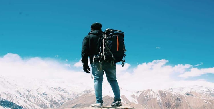 Backpacker standing on mountain peak with snowy slopes and blue sky, enjoying outdoor adventure.