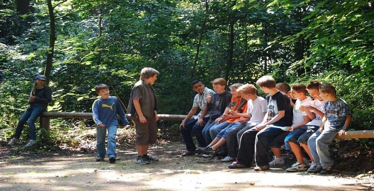 Young kids sitting outdoors on a forest bench during a reading activity.