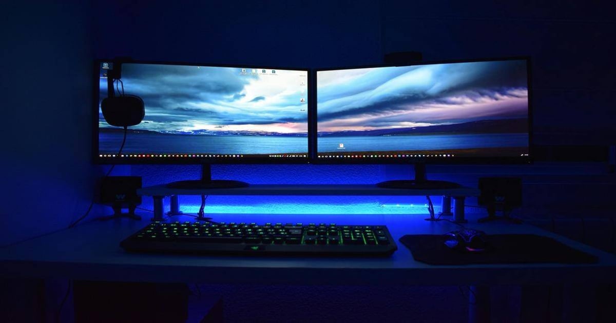 Computer desk with dual screens, gaming keyboard, and ambient blue LED backlit setup.