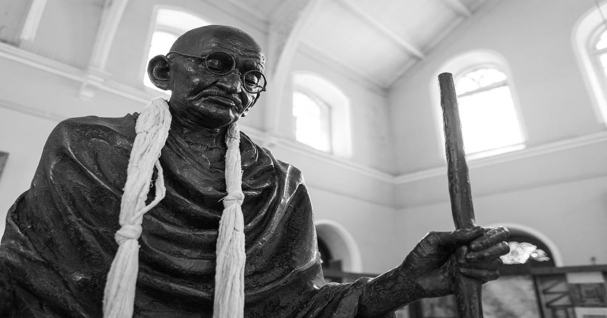 Mahatma Gandhi statue holding a walking stick, located inside a museum with large windows and high ceilings.