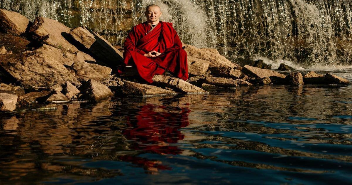 Meditating monk in red robes near waterfall with calm water reflection.