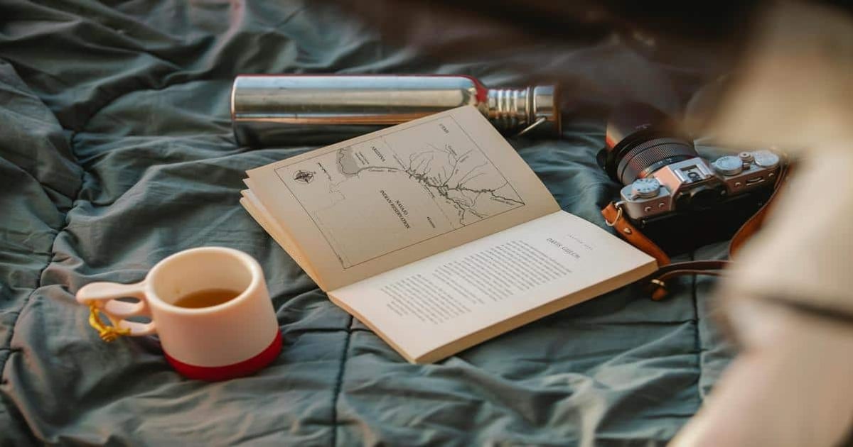 Portable camera, book, cup, and flashlight on bed for writing and reading.