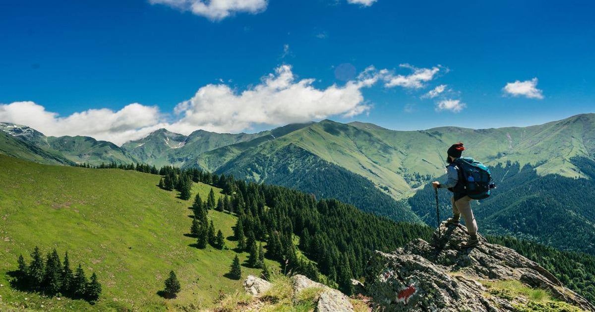 Hiker with backpack on rocky mountain ledge overlooking lush green hills and mountain range under blue sky.
