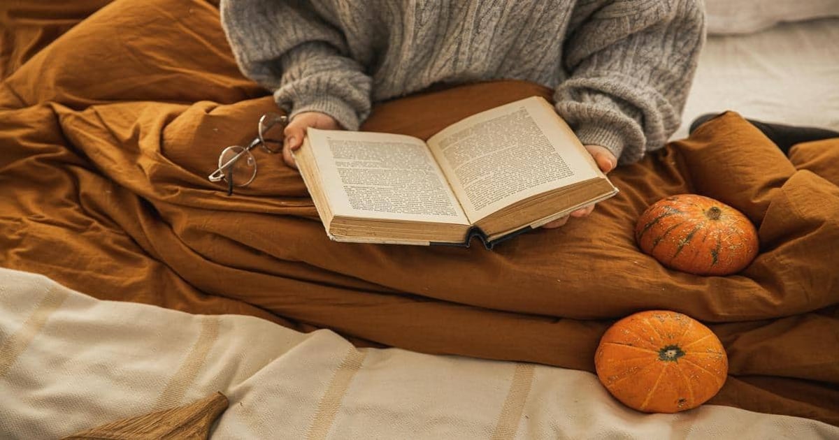 Close-up of person reading a book on bed with pumpkins for autumn vibe.