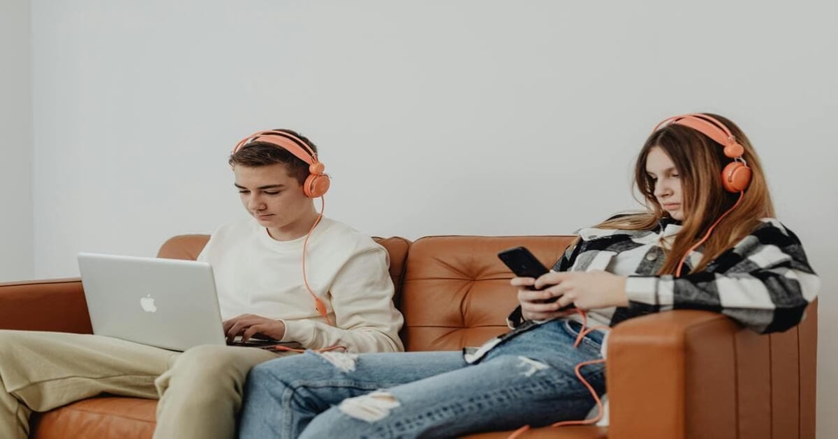 Alt text: Two teenagers with headphones using laptop and smartphone on brown couch, focusing on word and character counting.