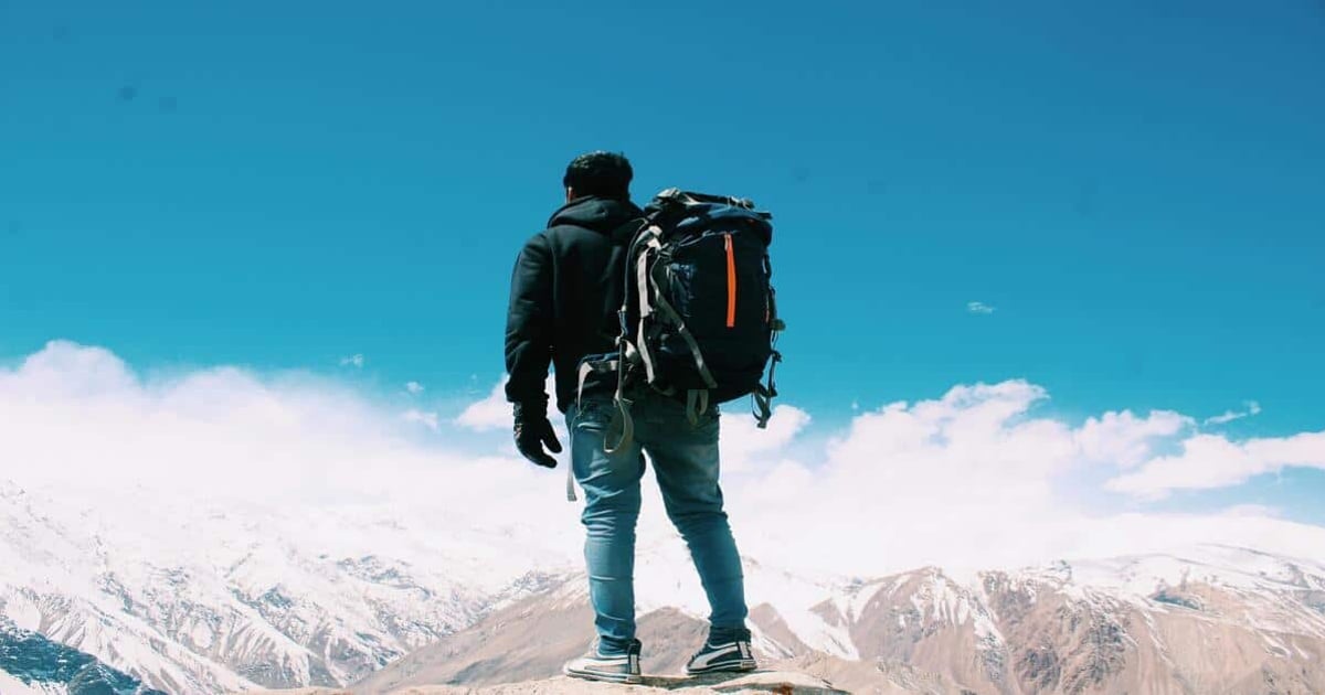 Backpacker standing on mountain peak with snowy slopes and blue sky, enjoying outdoor adventure.